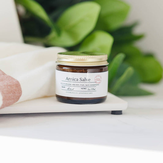 Jar of Arnica Salve on a white surface with green leaves in the background