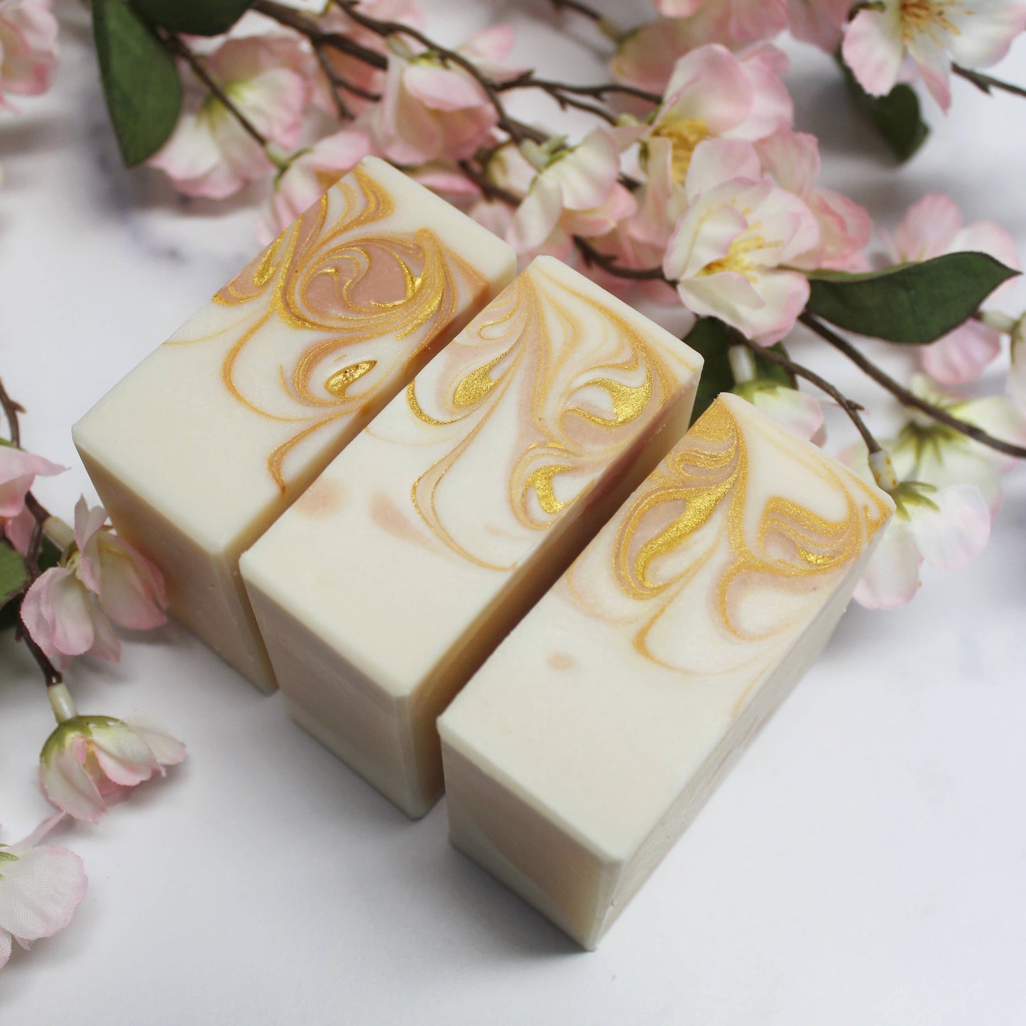 Three bars of soap with gold swirles on a white surface with pink flowers in the background