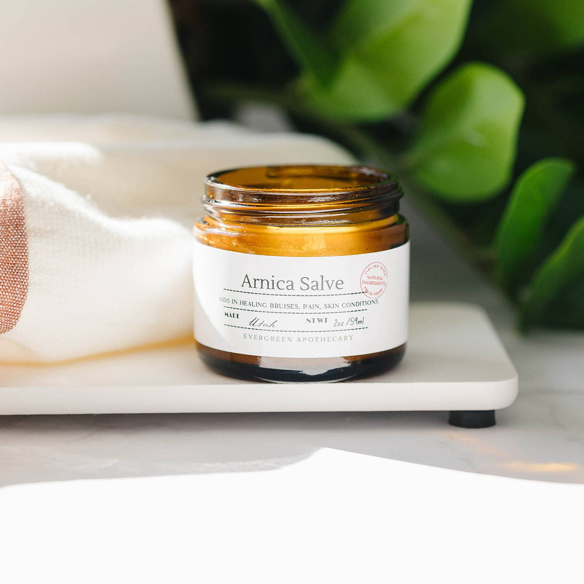 Jar of Arnica Salve on a marble surface with a blurred green plant in the background