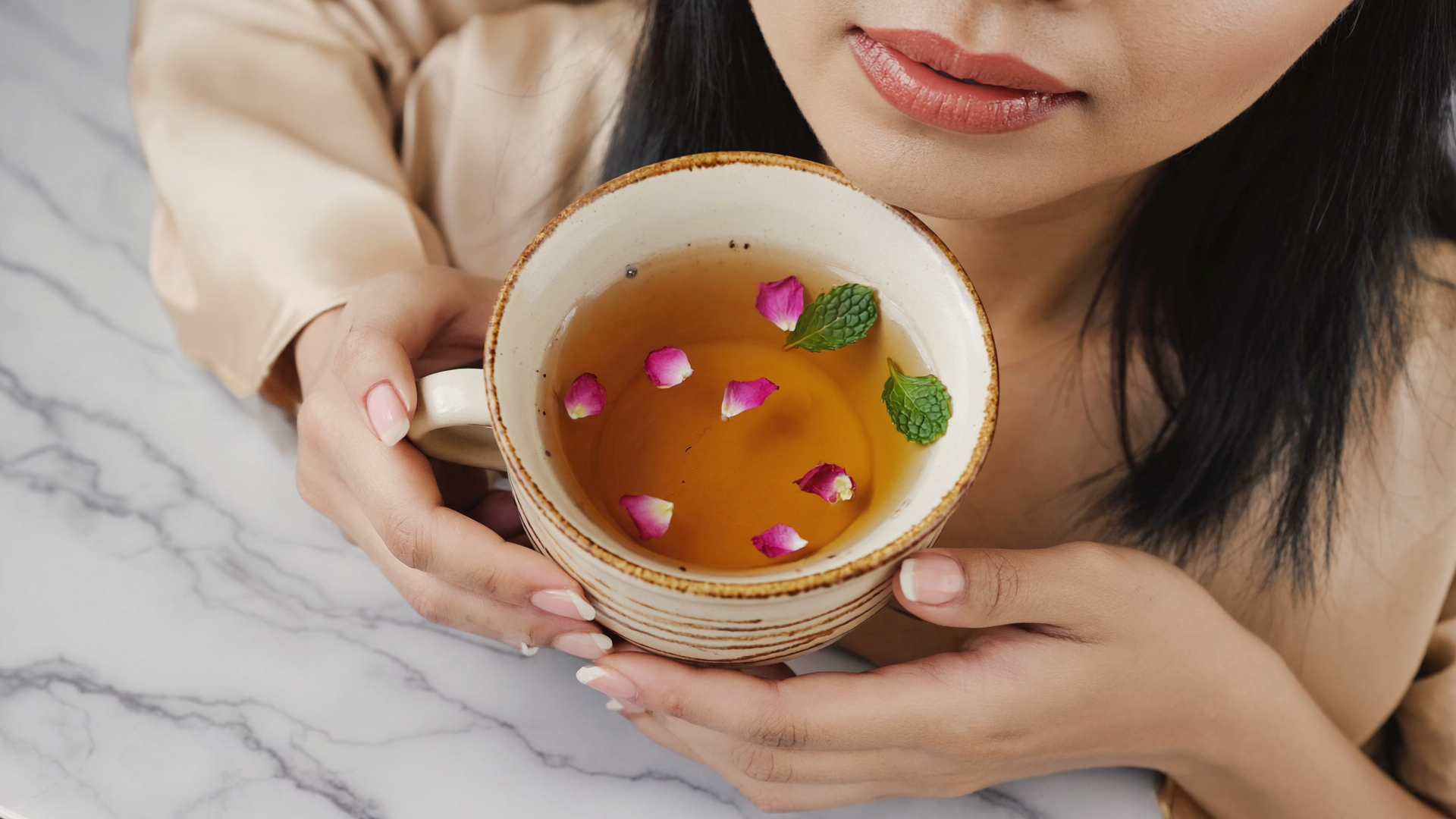 Woman holding a cup of tea with rose petals and mint leaves on a marble surface