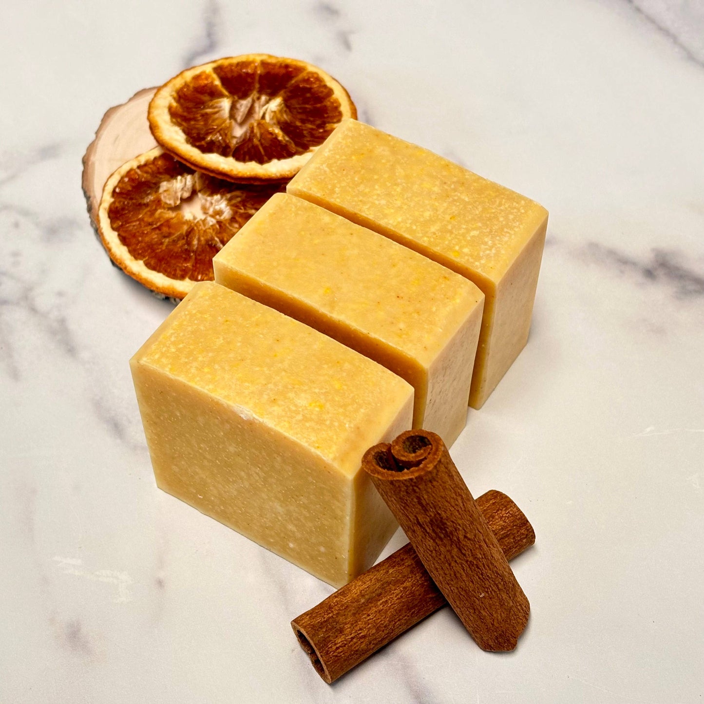 Three square bars of soap with orange slices and cinnamon sticks on a marble surface