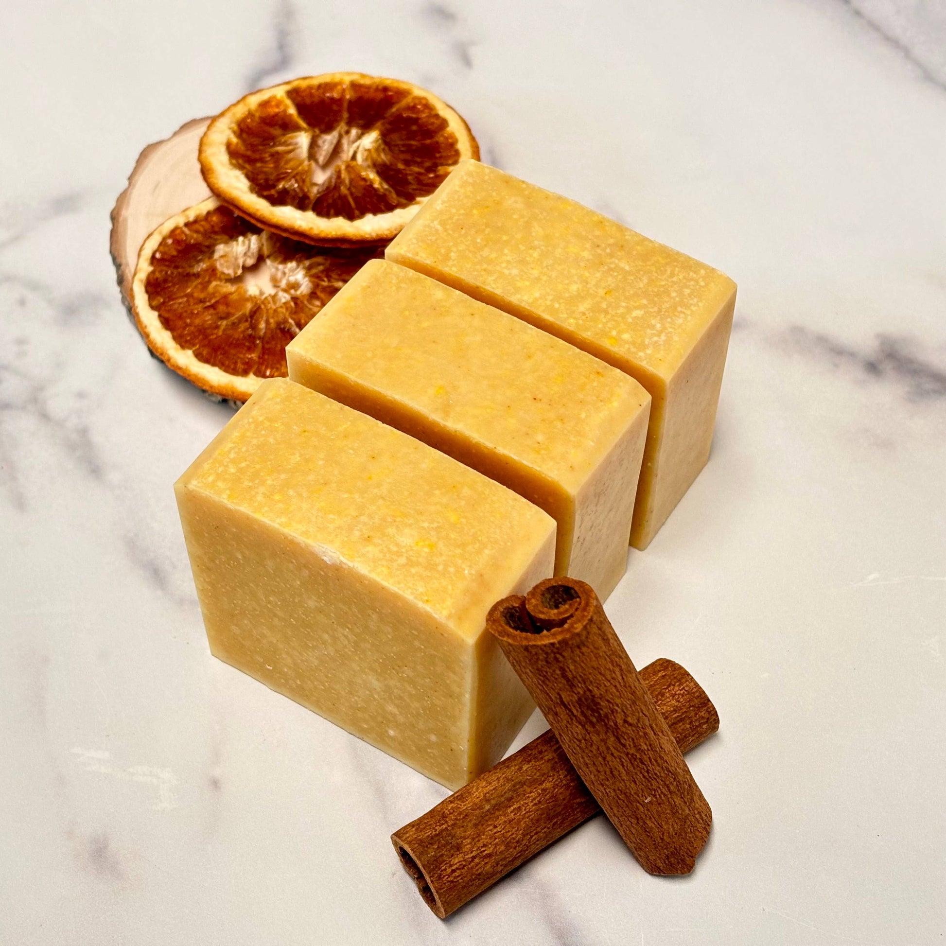 Three square bars of soap with orange slices and cinnamon sticks on a marble surface