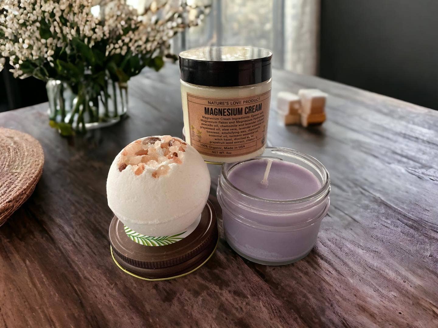 Bath bomb, jar of magnesium cream, and candle on a wooden surface with flowers in the background.