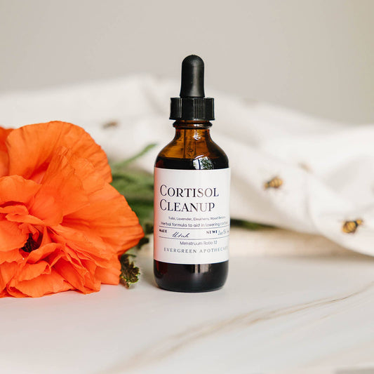An amber glass dropper bottle labeled 'CORTISOL CLEANUP' by Evergreen Apothecary, set on a white marble surface next to a bright orange flower, with a soft white fabric backdrop.
