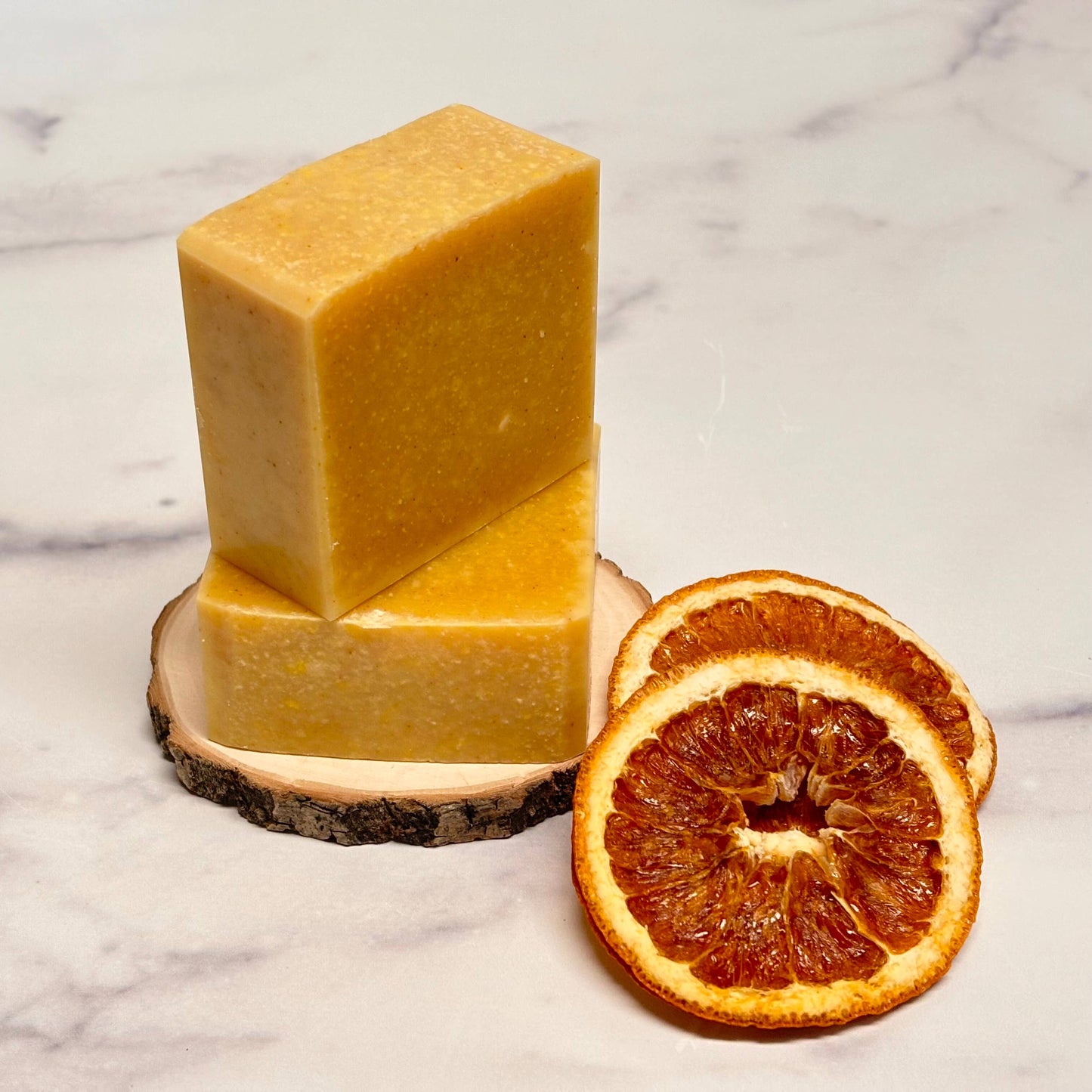 Two bars of yellow soap on a wooden block with dried orange slices on a marble background