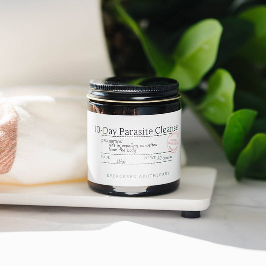 A glass supplement bottle labeled '10-Day Parasite Cleanse' by Evergreen Apothecary, resting on a white marble tray with a white cloth and green leaves in the background.