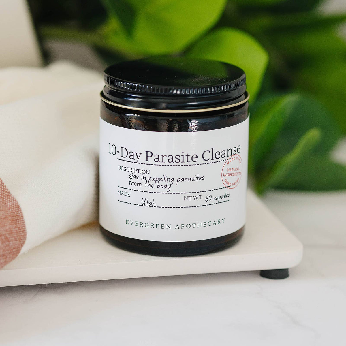 Jar of '10-Day Parasite Cleanse' by Evergreen Apothecary on a white surface with greenery in the background