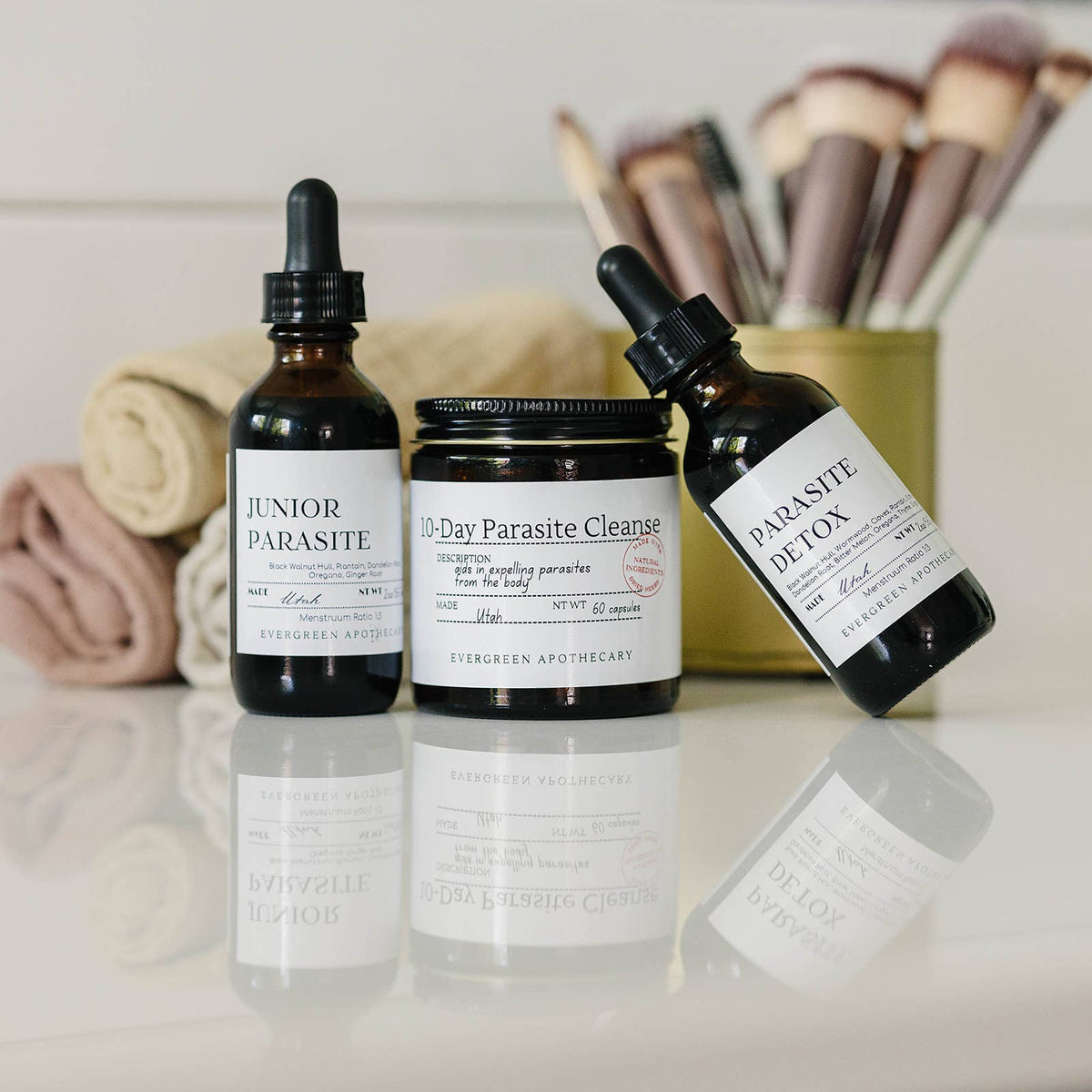 Three bottles of 'Parasite Detox' products on a reflective surface with makeup brushes in the background.
