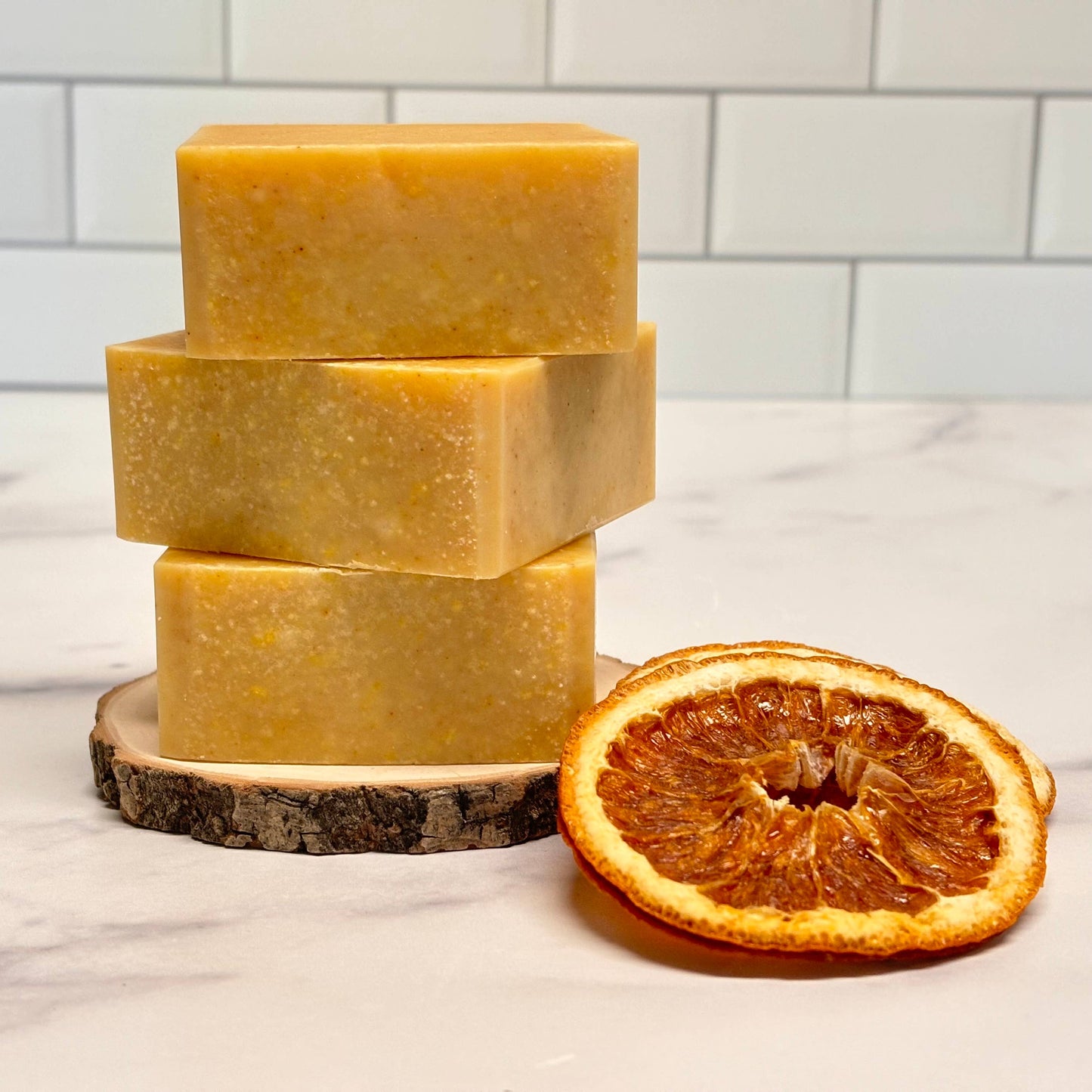 Three bars of yellow soap stacked on a wooden block with a dried orange slice beside them, against a white tiled wall.
