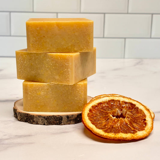 Three bars of yellow soap stacked on a wooden block with a dried orange slice beside them, against a white tiled wall.